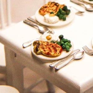 May include: Miniature plates of food, including grilled meat, green vegetables, and a white side dish, are arranged on a white table. Silverware, including forks and spoons, is placed around the plates. The scene suggests a small-scale dining experience.