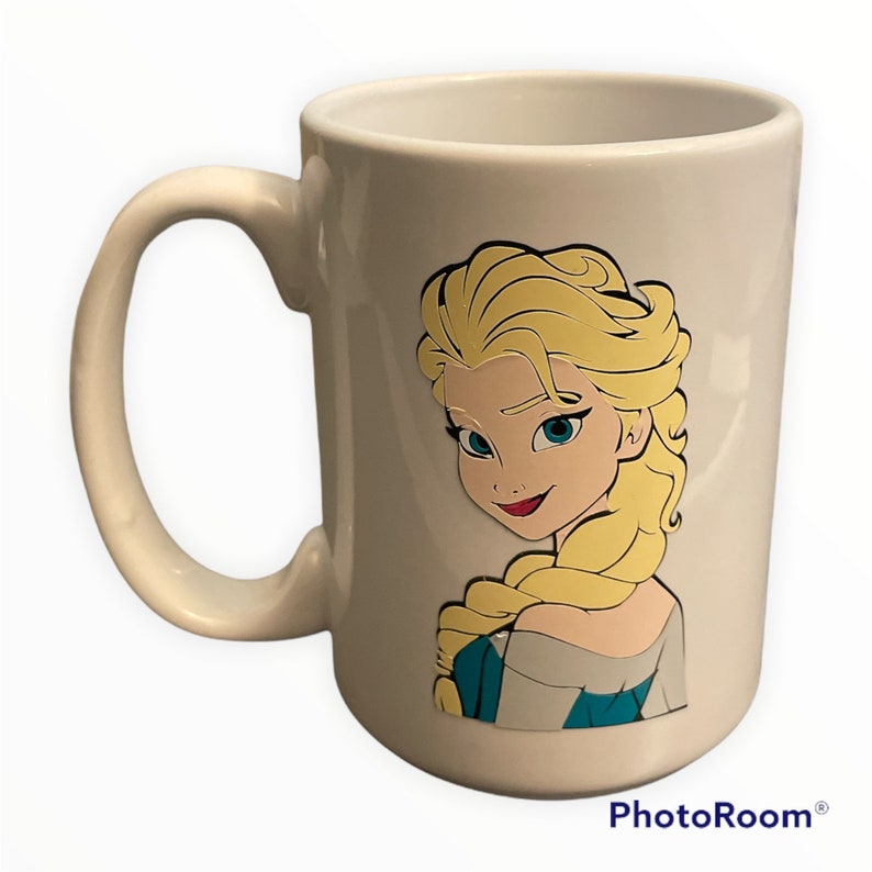 May include: White ceramic mug featuring a colourful cartoon image of Elsa from the film Frozen. The image shows Elsa looking over her shoulder with her long blonde hair.