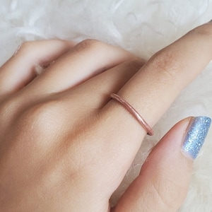 May include: A slender, rose gold ring is worn on a finger. The ring has a simple, minimalist design. The nail polish on the adjacent finger is a light blue with glitter.
