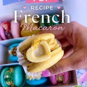 May include: A box of colorful French macarons, with one held up to the camera. The macaron in focus is yellow with a heart-shaped filling. The box contains macarons in various colors, including pink, blue, and yellow. Text on the image reads "PDF Recipe French Macaron."