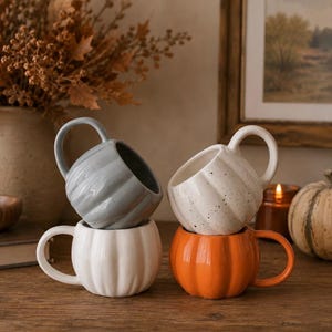 Könnte beinhalten: Ein Set aus vier kürbisförmigen Keramiktassen mit gerippter Textur. Die Kollektion umfasst eine glänzend graue Tasse, eine einfarbig weiße Tasse, eine cremefarbene Tasse mit dunklen Sprenkeln und eine leuchtend orangefarbene Tasse, jeweils mit Henkel.