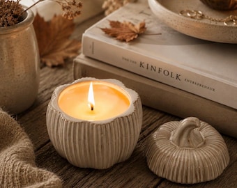 Op de afbeelding: Een brandende kaars in een crèmekleurige, geribbelde, pompoenvormige keramische pot met een bijpassend deksel ernaast. De kaars heeft een warm gloeiende vlam. De keramische houder heeft een textuur met verticale ribbels en een deksel met een steeltje.