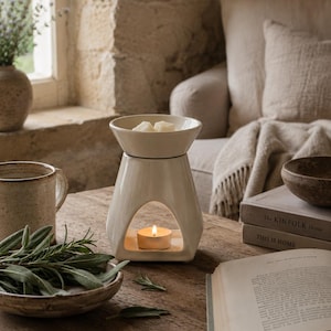 May include: A cream-colored ceramic wax warmer with a flared top bowl containing two white wax melts. The base features a triangular cutout housing a lit tea light candle. The warmer has a smooth, glossy finish and a wide, stable square-shaped bottom.