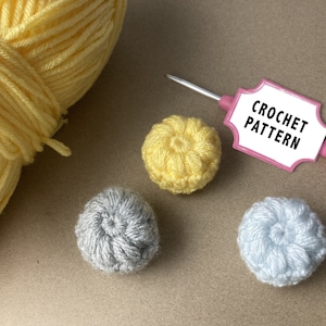 Puede incluir: Primer plano de suministros de ganchillo. Una madeja de hilo amarillo está junto a tres pequeñas esferas de ganchillo en amarillo, gris y azul claro. Un ganchillo rosa sostiene una etiqueta que dice "CROCHET PATTERN".