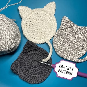 May include: Three crocheted cat coasters in gray and white yarn. The coasters are shaped like cats with tails. A crochet hook and a sign that says "Crochet Pattern" are also in the image.