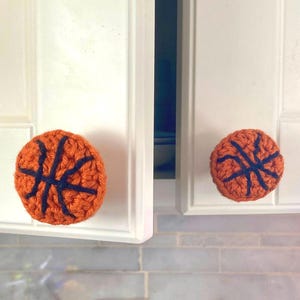 Peut inclure: Deux boutons de placard en crochet orange et bleu marine en forme de ballon de basket, fixés à des portes de placard blanches. Les boutons sont ronds et présentent le design classique du ballon de basket. Les portes sont fermées.