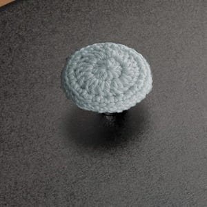 May include: A pale blue, crocheted knob, circular in form, is positioned on a dark grey surface. The knob is crafted from yarn and has a textured finish. The background is a solid, dark grey colour.