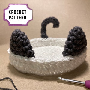 Könnte beinhalten: Häkelmuster für einen weißen und grauen Katzenkorb. Der Korb hat zwei graue Ohren, einen grauen Schwanz und einen weißen Körper. Der Korb steht auf einer beigefarbenen Oberfläche.