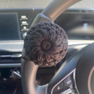 Może przedstawiać: Zbliżenie na ciemnobrązową, szydełkową osłonę kierownicy, o kulistym kształcie. Osłona wykonana jest z teksturowanej przędzy i umieszczona na czarnej kierownicy. Tło przedstawia wnętrze samochodu.