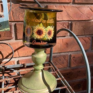 May include: A decorative candle holder with a green base and a clear glass top. The glass is printed with sunflowers and the words "Spread a little Sunshine". The candle holder is displayed on a metal shelf against a brick wall.