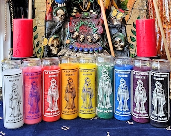May include: A collection of eight colorful candles with white lettering that reads "La Santisima Muerte" and "Holy Death". The candles are arranged in a row on a black surface in front of a religious altar with a skeletal figure and other religious imagery.
