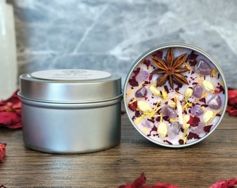 May include: A silver tin candle with a pink wax top. The wax is filled with dried flower petals, amethyst crystals, and a star anise. The candle is in a silver tin with a white label.