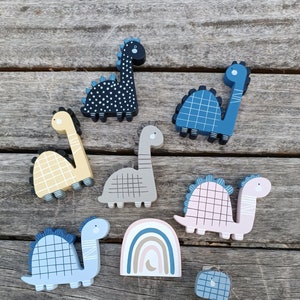 Peut inclure: Un ensemble de huit figurines de dinosaures en bois de différentes couleurs et motifs, notamment bleu, rose, jaune, noir et gris. Les dinosaures sont disposés sur une surface en bois avec une découpe en bois en forme d'arc-en-ciel.