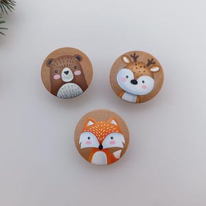 以下が含まれることがあります： クマ、シカ、キツネの顔が描かれた3つの木製キャビネットノブ。クマは茶色で白い胸、シカはタン色で角があり、キツネはオレンジと白です。ノブは白い表面に配置されています。
