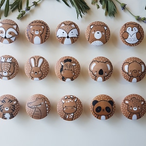 Op de afbeelding: Een verzameling van vijftien houten kastknoppen, elk met een handgeschilderd dierenontwerp. De knoppen zijn bruin met witte stippen. De dieren zijn onder meer een wasbeer, egel, vos, beer, uil, hert, konijn, zebra, koala, olifant, tijger, alligator, giraffe, panda en luipaard.