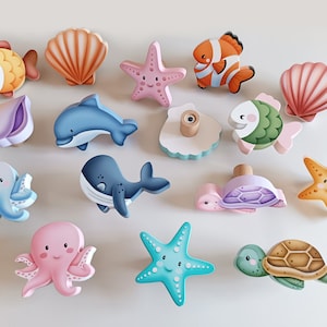 Op de afbeelding: Een set van 12 houten deurknopjes met ocean-thema ontwerpen. De knopjes hebben verschillende zeedieren, waaronder een ster, een zeepaardje, een dolfijn, een walvis, een octopus, een schildpad, een schelp en een vis. De knopjes zijn geschilderd in heldere, vrolijke kleuren.
