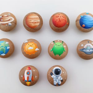 May include: A collection of ten wooden cabinet knobs with colorful space-themed designs. Each knob features a different planet, the sun, a green alien, a spaceship, a rocket, and an astronaut. The knobs are round and made of light-colored wood.