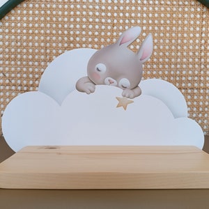 May include: A white wooden shelf shaped like a cloud with a grey bunny rabbit sleeping on it. The bunny is holding a small yellow star. The shelf is on a brown surface.