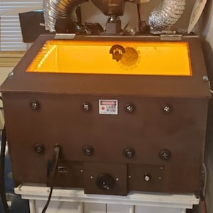 Peut inclure: Une boîte en métal marron avec un couvercle transparent et une lumière orange vive à l'intérieur. La boîte porte une pancarte d'avertissement indiquant "DANGER LASER EN SERVICE".