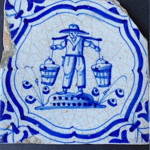 May include: A blue and white Delft tile with a decorative border and a central image of a person carrying two buckets on a yoke.