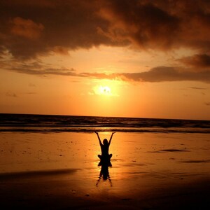 Puede incluir: Silueta de una persona sentada en una playa con los brazos levantados hacia una vibrante puesta de sol naranja. El cielo está lleno de nubes y las olas del océano son visibles en la distancia. La escena se refleja en la arena mojada.