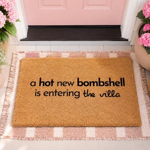 May include: A rectangular tan coir doormat featuring the black text "a hot new bombshell is entering the villa" centered on the surface. The mat is positioned on a light pink and white striped rug in front of a light pink door with white trim.