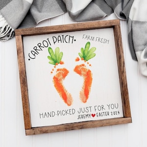 May include: A square wooden sign with a white background featuring two orange footprint-shaped carrots with green tops. The text reads: CARROT PATCH. FARM FRESH. HAND-PICKED JUST FOR YOU. JEREMY [heart symbol] EASTER 2025. The sign has a dark wood frame.