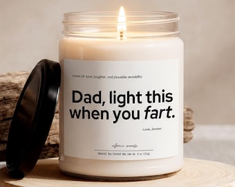 May include: A lit candle in a clear glass jar with a black lid. The candle has a white label with the text "Dad, light this when you fart." The candle sits on a wooden disc.