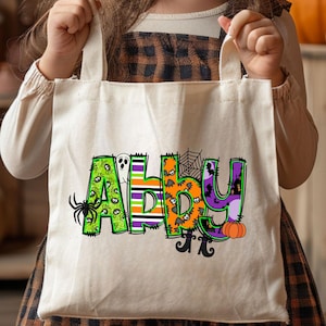 May include: A Halloween-themed tote bag with the name Abby in colorful, decorated letters. The letters are decorated with various Halloween elements such as spiders, ghosts, pumpkins, and bats. The bag is off-white canvas and has a simple design.