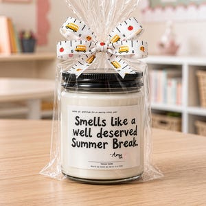 May include: A candle in a glass jar with a black lid, wrapped in clear cellophane and tied with a ribbon. The ribbon features school buses and apples. The label reads "Smells like a well deserved Summer Break."