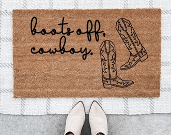 Può includere: Un tappetino da porta marrone con il testo "boots off, cowboy." in lettere nere. Due stivali da cowboy neri sono illustrati sul tappetino.