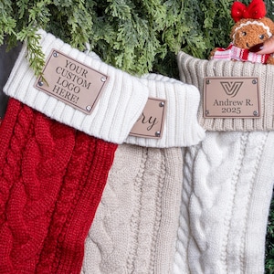 May include: Three cable-knit Christmas stockings in red, beige, and white hang from a garland. Each stocking has a white cuff and a brown leather tag with custom text. A gingerbread man with a red bow is placed in one stocking.