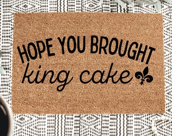 Może przedstawiać: Prostokątna wycieraczka z włókna kokosowego w kolorze brązowym z czarnym napisem HOPE YOU BROUGHT king cake oraz symbolem fleur-de-lis. Mata ma szorstką, naturalną strukturę włókien i jest ułożona poziomo z wyraźnym, czarnym nadrukiem na środku.