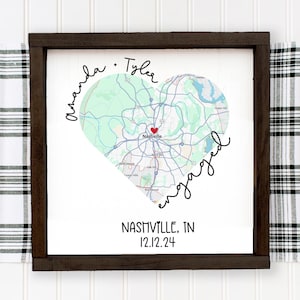 May include: A white framed sign with a map of Nashville, Tennessee in the shape of a heart. The text "Amanda - Tyler engaged" is written around the heart. The text "Nashville, TN 12.12.24" is written below the heart.
