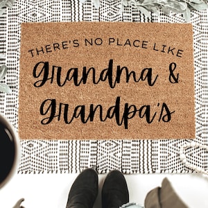 May include: A rectangular coir doormat with the text "THERE'S NO PLACE LIKE grandma & grandpa's" in black script. The mat is placed on a patterned rug. A coffee cup and boots are visible in the frame, adding context to the scene.