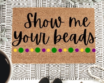 Może przedstawiać: Prostokątna wycieraczka z naturalnego brązowego włókna kokosowego. Mata zawiera napis "Show me your beads" czarnym skryptem, z rzędem żółtych, zielonych i fioletowych kropek. Mata jest umieszczona na wzorzystym dywanie.