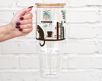 May include: A tall clear glass tumbler with a handle, featuring a bamboo lid and a clear straw. The exterior is decorated with a graphic print of a black cat, potted plants, books, and a coffee cup in shades of green, brown, and soft pink.