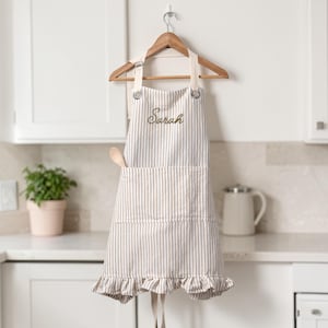 May include: A beige and white striped apron featuring a ruffled hem, a front pocket, and the name Sarah embroidered in brown thread on the chest. The apron is suspended from a wooden hanger by cream-colored fabric straps with metal grommets.