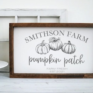 May include: A rectangular wooden sign with a dark brown frame and a white background. The sign features gray text that reads SMITHSON FARM, pumpkin patch, FARM FRESH, and EST. 2021, along with a detailed line drawing of three pumpkins.
