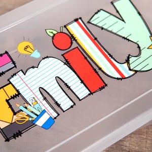 Może przedstawiać: P&oacute;łprzezroczysty plastikowy pi&oacute;rnik z nadrukiem imienia EMILY w dużych, kolorowych literach o tematyce szkolnej. Wz&oacute;r zawiera ż&oacute;łtą żar&oacute;wkę, czerwone jabłko, ż&oacute;łtą gwiazdkę oraz ilustracje oł&oacute;wk&oacute;w, nożyczek i papieru.