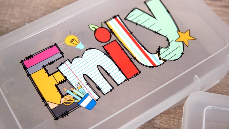 Może przedstawiać: P&oacute;łprzezroczysty plastikowy pi&oacute;rnik z nadrukiem imienia EMILY w dużych, kolorowych literach o tematyce szkolnej. Wz&oacute;r zawiera ż&oacute;łtą żar&oacute;wkę, czerwone jabłko, ż&oacute;łtą gwiazdkę oraz ilustracje oł&oacute;wk&oacute;w, nożyczek i papieru.