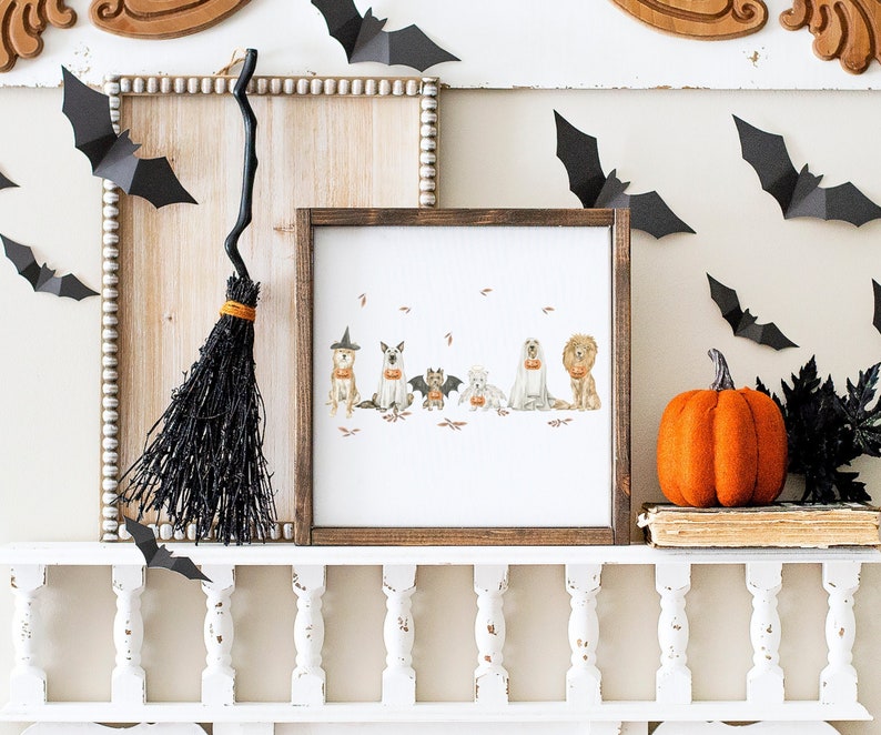 May include: A framed print of a group of dogs dressed in costumes holding pumpkins. The print is on a white background and is surrounded by a brown frame. The print is hanging on a white mantel with a black broom and a pumpkin on the shelf below.