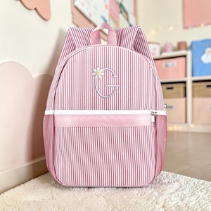 May include: A pink and white striped backpack featuring a top handle, two shoulder straps, and a front zippered pocket. The center of the backpack is embroidered with a large light gray letter G accented by a small white daisy flower detail.