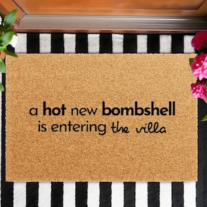 May include: A rectangular tan coir doormat with black text that reads, a hot new bombshell is entering the villa. The mat has a coarse, natural fiber texture and is positioned on a black and white striped surface with potted flowers on the sides.