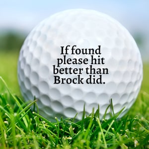 May include: A white golf ball with a dimpled surface featuring the black printed text: If found please hit better than Brock did. The golf ball is positioned in a patch of green grass, with the focus centred on the text printed on the ball.
