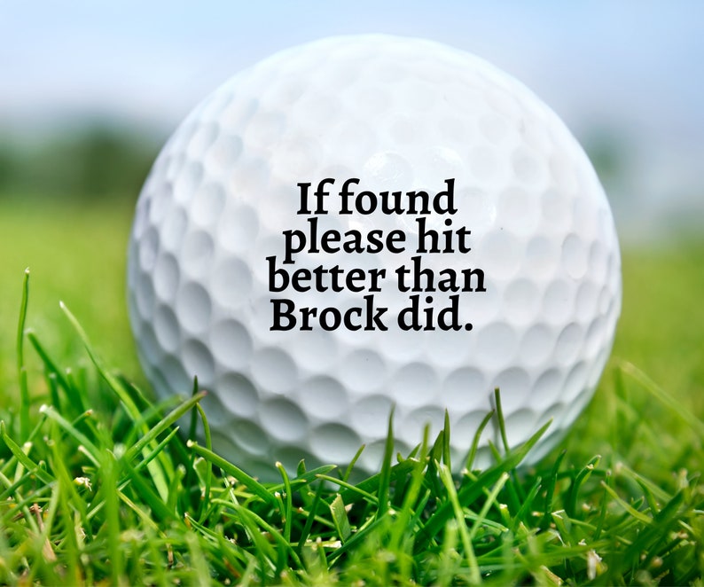 May include: A white golf ball with a dimpled surface featuring the black printed text: If found please hit better than Brock did. The golf ball is positioned in a patch of green grass, with the focus centred on the text printed on the ball.