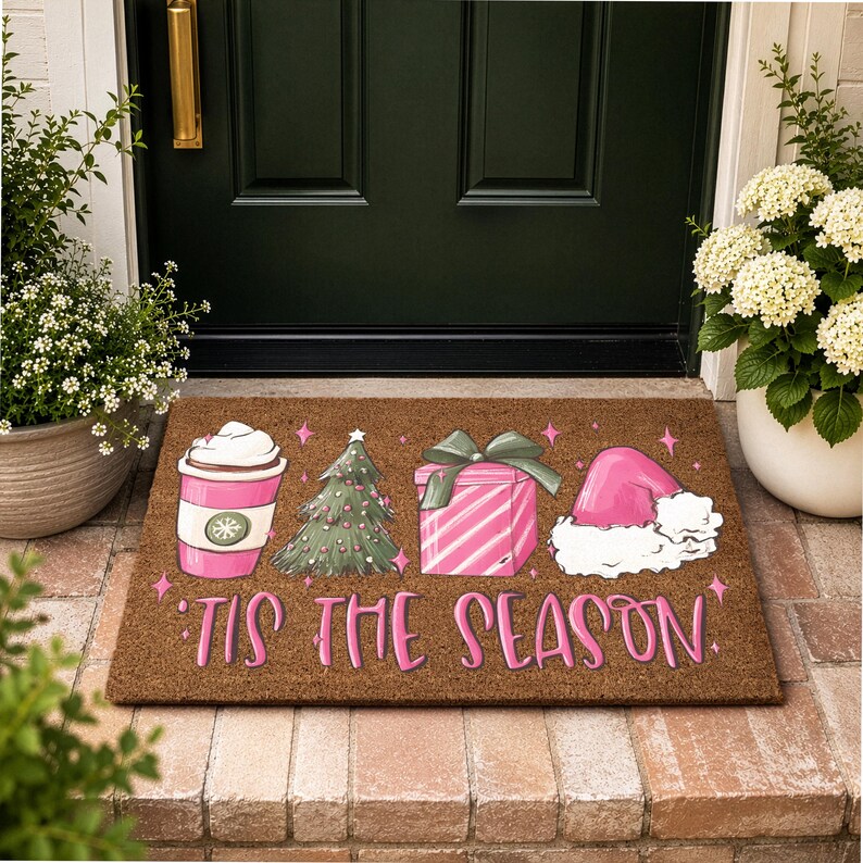 Op de afbeelding: Een rechthoekige bruine kokosmat met een roze koffiebeker, een groene kerstboom, een roze cadeau met een groene strik en een roze kerstmuts. De tekst 'TIS THE SEASON' staat in roze letters onder de illustraties met kleine roze sterretjes.