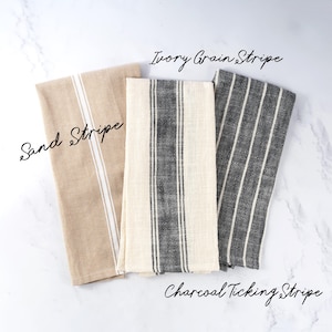 May include: Three rectangular kitchen towels featuring striped patterns. The left towel is tan with a white stripe labeled Sand Stripe. The center towel is ivory with a dark gray stripe labeled Ivory Grain Stripe. The right towel is dark gray with white stripes labeled Charcoal Ticking Stripe.