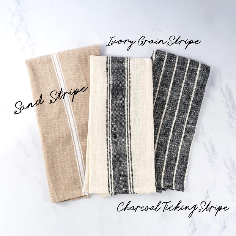 May include: Three rectangular kitchen towels featuring striped patterns. The left towel is tan with a white stripe labeled Sand Stripe. The center towel is ivory with a dark gray stripe labeled Ivory Grain Stripe. The right towel is dark gray with white stripes labeled Charcoal Ticking Stripe.