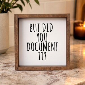 Brown-framed “But Did You Document It?” sign on a marble counter with a plant and candle in the background.